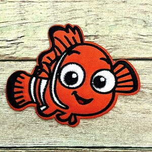 May include: An orange embroidered patch in the shape of a cartoon clownfish. The fish has large white eyes with black pupils, black outlines, and black and white stripes. The patch is on a light brown wooden surface.