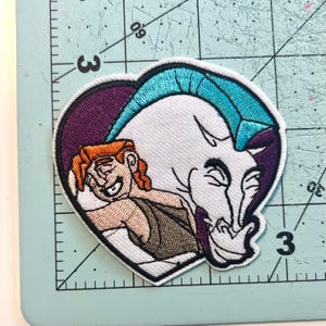 May include: Embroidered patch featuring Hercules and Pegasus from the Disney movie. The patch is heart-shaped with a purple border. Hercules has orange hair and is smiling. Pegasus is white with a turquoise mane and a purple outline.
