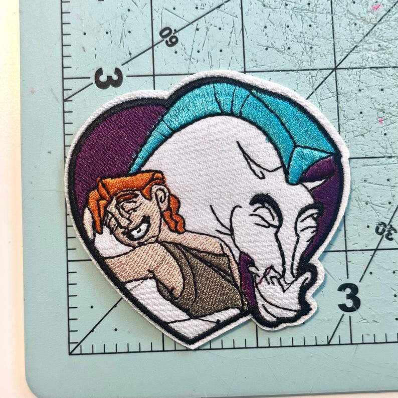 May include: Embroidered patch featuring Hercules and Pegasus from the Disney movie. The patch is heart-shaped with a purple border. Hercules has orange hair and is smiling. Pegasus is white with a turquoise mane and a purple outline.