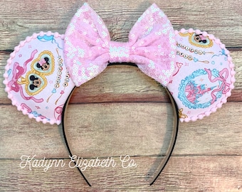 May include: Pink sequined bow and mouse ears headband. The ears are decorated with a pink and white pattern featuring Mickey Mouse and a castle. The headband is black, and the text "Kadynn Elizabeth Co" is visible.