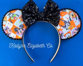 May include: Black sequined Minnie Mouse ears with a Halloween-themed fabric featuring various Disney characters, including Mickey Mouse, Minnie Mouse, Goofy, Donald Duck, and Pluto. The fabric is orange, white, and black with a checkered pattern. The text "Kadyrn Elizabeth Co." is printed on the headband.