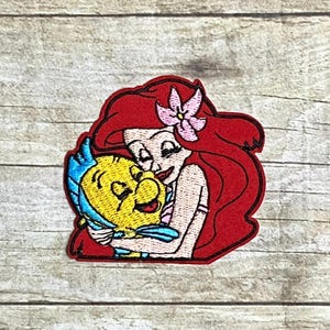 May include: Embroidered patch featuring Ariel from The Little Mermaid hugging Flounder. Ariel has red hair and a pink flower. Flounder is yellow with blue fins. The patch has a red border.