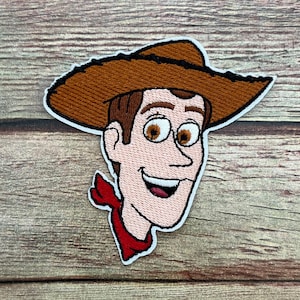 May include: Embroidered patch featuring a cartoon cowboy character with a brown hat, hair, and eyes. The character has a red neckerchief and a smiling expression. The patch has a white border.