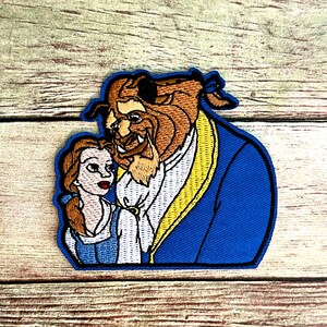 May include: Embroidered patch featuring the Beast and Belle from Beauty and the Beast. The Beast is depicted with brown fur and a blue coat, while Belle wears a blue and white dress. The patch has a blue border.