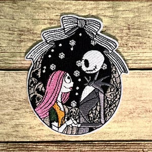 May include: Embroidered patch featuring Jack Skellington and Sally from The Nightmare Before Christmas. The patch is shaped like an ornament with a black background, snowflakes, and a white bow at the top. Sally has pink hair and a yellow dress.