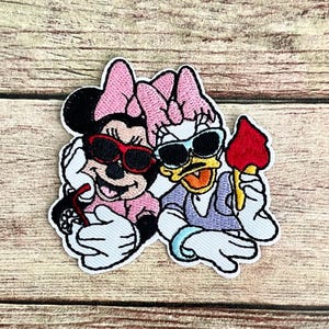 May include: Embroidered patch featuring Minnie Mouse and Daisy Duck wearing sunglasses. Minnie Mouse is holding a drink with a straw, and Daisy Duck is holding a red ice cream cone. Both characters are wearing pink bows and smiling.