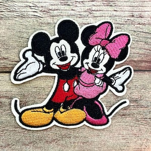 May include: Embroidered patch featuring Mickey Mouse and Minnie Mouse. Mickey is wearing red shorts and yellow shoes, while Minnie is in a pink dress and bow. The patch has a white border and is set against a wood grain background.