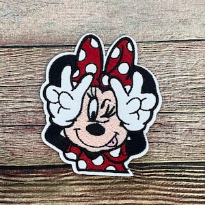 May include: Embroidered patch featuring a cartoon character with a red bow with white polka dots. The character is smiling and making a peace sign with both hands. The patch has a white border and is set against a wooden background.