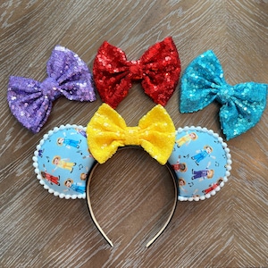 May include: A pair of blue fabric mouse ears with a white trim and a pattern of cartoon characters. The ears are decorated with four different colored bows: purple, red, yellow, and blue.