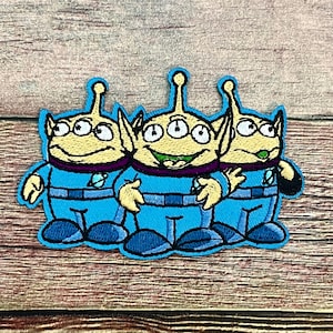 May include: Embroidered patch featuring three cartoon aliens with three eyes, wearing blue suits with purple accents. The aliens have a smiling expression and are outlined in blue.