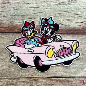 May include: Embroidered patch featuring Daisy Duck and Minnie Mouse in a pink convertible car. Daisy Duck is wearing a blue top and red sunglasses. Minnie Mouse is wearing a red top and a blue bow. The car has yellow headlights and pink wheels.