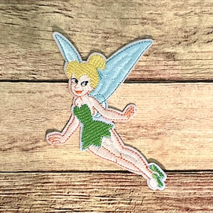 May include: Embroidered patch featuring a cartoon fairy with blonde hair, light blue wings, and a green dress. The fairy is in a flying pose, with a smiling expression. The patch has a white border and is on a wooden background.