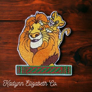 May include: Embroidered patch featuring a smiling lion with a cub on its head. The lion has a large, orange mane and a friendly expression. The patch includes a green and red rectangular border and the text "Kadynn Elizabeth Co."