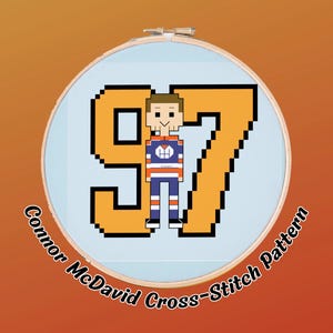 May include: A cross-stitch pattern featuring a pixelated image of a hockey player in a blue, orange, and white uniform, standing in front of the number 97. The text "Connor McDavid Cross-Stitch Pattern" is in a circular design.