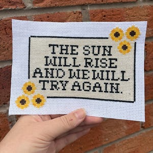 May include: Cross-stitch art piece with the text "THE SUN WILL RISE AND WE WILL TRY AGAIN." The design features a cream-colored background with black lettering and small yellow sunflower accents. The piece is held against a brick wall.
