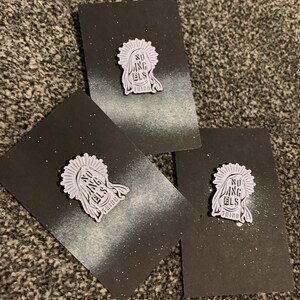 Può includere: Tre carte nere con macchie bianche, ciascuna con uno spillo in smalto viola con un'immagine stilizzata di una testa di persona con il testo "NO ANGELS THIRD" su di essa.