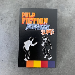 Könnte beinhalten: Schwarzer Emaille-Pin mit dem Text "PULP FICTION Jackrabbit Slims" in Orange und Blau. Zwei Figuren in Schwarz und Weiß tanzen auf dem Pin. Ein farbenfroher Streifen aus Orange, Gelb, Rot und Lila befindet sich am unteren Rand.