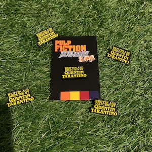 May include: A black card with the text "Pulp Fiction Jackrabbit Slims" and four yellow and black pins with the text "Written and Directed by Quentin Tarantino" are displayed on a bed of green grass.