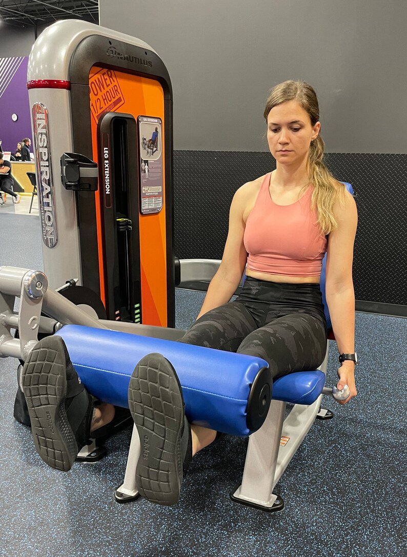 May include: A woman is using a leg extension machine at the gym. The machine is silver and blue with the words "Inspiration" and "Leg Extension" printed on it. The machine has a blue padded seat and a blue padded footrest. The woman is wearing a pink sports bra and black leggings.