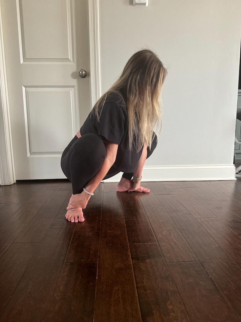 May include: A person wearing black leggings and a black short-sleeved shirt is performing a squat exercise on a wooden floor.