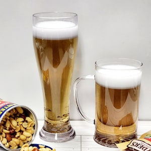 May include: Two clear glass beer mugs filled with light beer and a white head of foam. A can of peanuts is tipped over, spilling peanuts onto a white wooden surface. A bag of pretzels is on the surface next to the mugs.