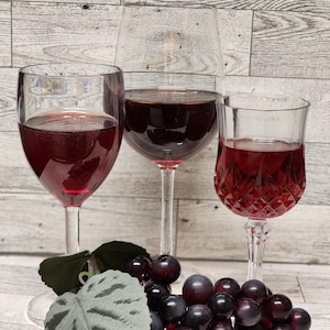 May include: Three glasses of red wine with a bunch of dark purple grapes and a green leaf.