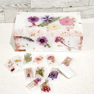 Peut inclure: Une boîte blanche avec un motif floral sur le couvercle et un jeu de dominos avec des fleurs pressées et des accents dorés.
