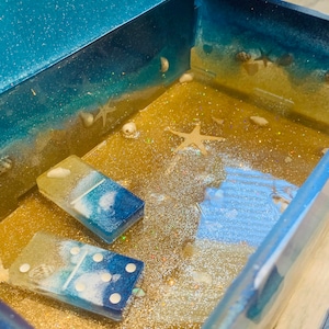 Könnte beinhalten: Eine blaue und goldene Schatztruhe mit einer Strandkulisse im Inneren. Die Truhe ist gefüllt mit Sand, Muscheln und zwei Dominosteinen. Die Dominosteine sind mit einem blauen und weißen Wellendesign bemalt.