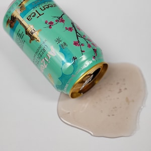 May include: A can of Arizona Green Tea with ginseng and honey, spilled on a white surface. The can is partially tipped over, with the green tea spilling out. The can has a green and blue design with pink flowers and the text "Arizona Green Tea with Ginseng and Honey" and "Real Brewed" printed on it.
