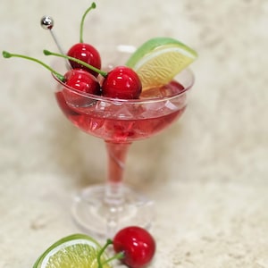 Puede incluir: Una copa de cóctel transparente con tallo llena de un líquido rojo y cubitos de hielo, decorada con tres cerezas rojas brillantes con tallo y una rodaja de lima. Dos cerezas adicionales y una rodaja de lima se sitúan frente a la base.
