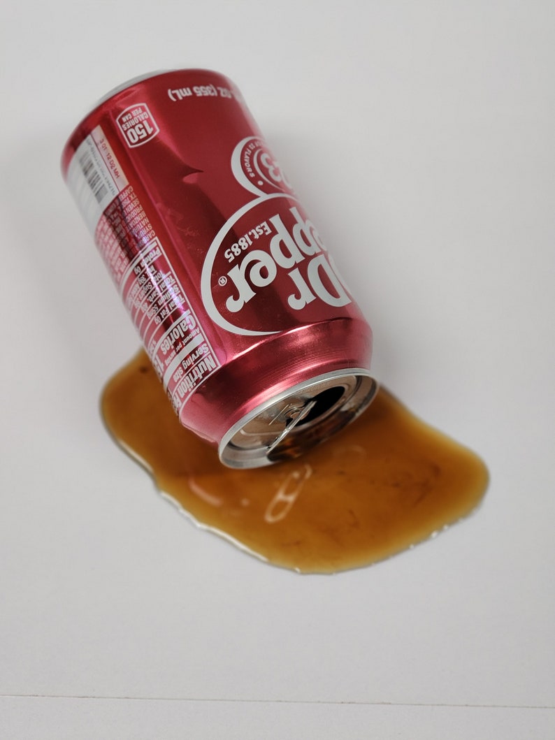 May include: A red aluminum can of Dr Pepper soda has spilled on a white surface, creating a brown puddle. The can is lying on its side with the Dr Pepper logo and text visible. The can is labeled "Est. 1885" and "355 mL (12 oz)".