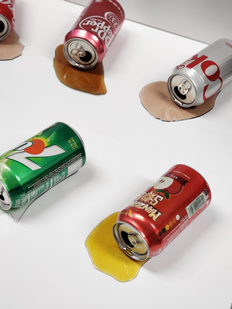 May include: Four aluminum cans of soda spilled on a white surface. The cans are red, green, silver, and orange. The red can says "Dr Pepper", the green can says "7 Up", the silver can says "Coke", and the orange can says "Mtn Dew".