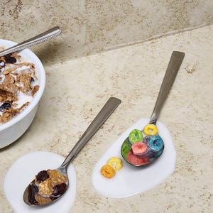 May include: Two spoons with cereal and milk on a speckled surface. One spoon holds a mix of cereal and raisins, the other holds colorful, ring-shaped cereal. A bowl of cereal is in the background.