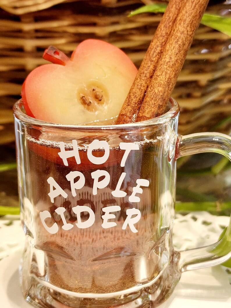 May include: A clear glass mug filled with hot apple cider, garnished with a cinnamon stick and a slice of apple. The words "HOT APPLE CIDER" are printed on the front of the mug. The drink is set against a blurred background.