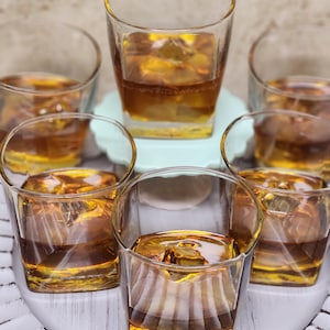 Puede incluir: Seis vasos transparentes de forma cuadrada llenos de líquido ámbar y cubitos de hielo. Los vasos están colocados sobre una bandeja blanca texturizada y un plato azul claro. Las bebidas parecen ser whisky o un licor similar.