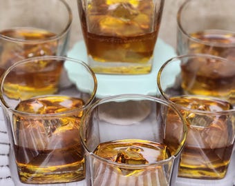 May include: Six clear, square-shaped glasses filled with amber liquid and ice cubes. The glasses are arranged on a white, textured tray and a light blue plate. The drinks appear to be whiskey or a similar spirit.