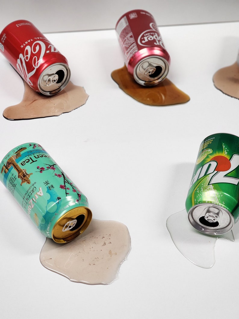 May include: Four aluminum beverage cans are lying on their sides on a white surface. The cans are all spilled and the liquid has pooled around them. The cans are Coca-Cola, Dr Pepper, Arizona Green Tea, and Mountain Dew.