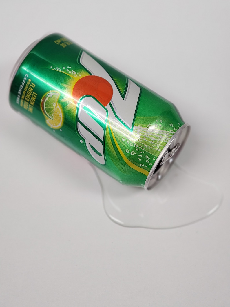 May include: A green and silver can of 7 Up lemon-lime soda with a lime slice and a red circle on the can. The can is lying on its side and there is a puddle of liquid on the white surface.