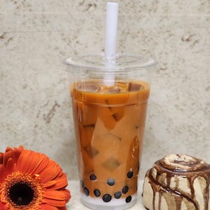 May include: A tall plastic cup of iced milk tea with boba pearls and a white straw. An orange gerbera daisy and a cinnamon roll are next to the drink. The drink is on a light-coloured surface.