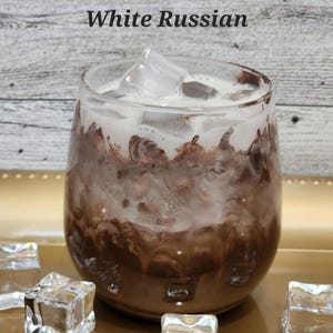 May include: A glass of White Russian cocktail with ice cubes on a gold tray. The drink is a creamy brown color with a white layer of cream on top.