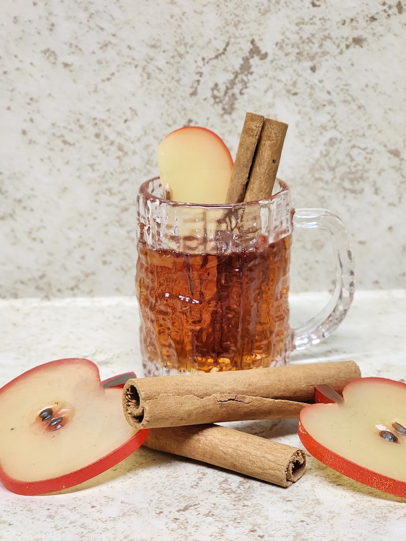 May include: A clear glass mug filled with a warm, amber-colored beverage, garnished with a slice of apple and two cinnamon sticks. Two apple halves and three cinnamon sticks are arranged in front of the mug, creating a cozy, autumnal scene.