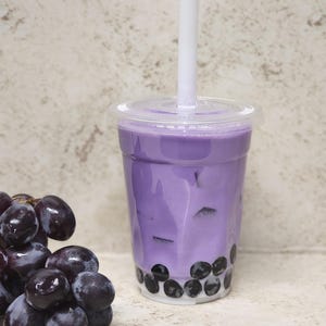 May include: A vibrant purple bubble tea in a clear plastic cup with a white straw. The drink contains black tapioca pearls. A cluster of dark purple grapes is positioned to the left of the drink.