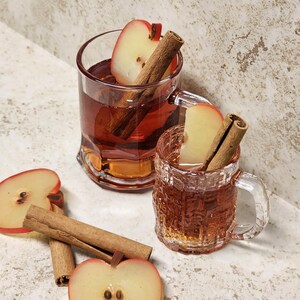 May include: Two clear glass mugs filled with a reddish-brown liquid, garnished with apple slices and cinnamon sticks. Slices of apples and cinnamon sticks are scattered around the mugs. The mugs have handles and textured sides, suggesting a warm beverage.