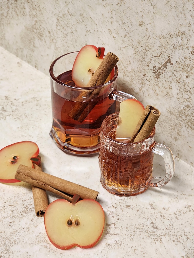 May include: Two clear glass mugs filled with a reddish-brown liquid, garnished with apple slices and cinnamon sticks. Slices of apples and cinnamon sticks are scattered around the mugs. The mugs have handles and textured sides, suggesting a warm beverage.