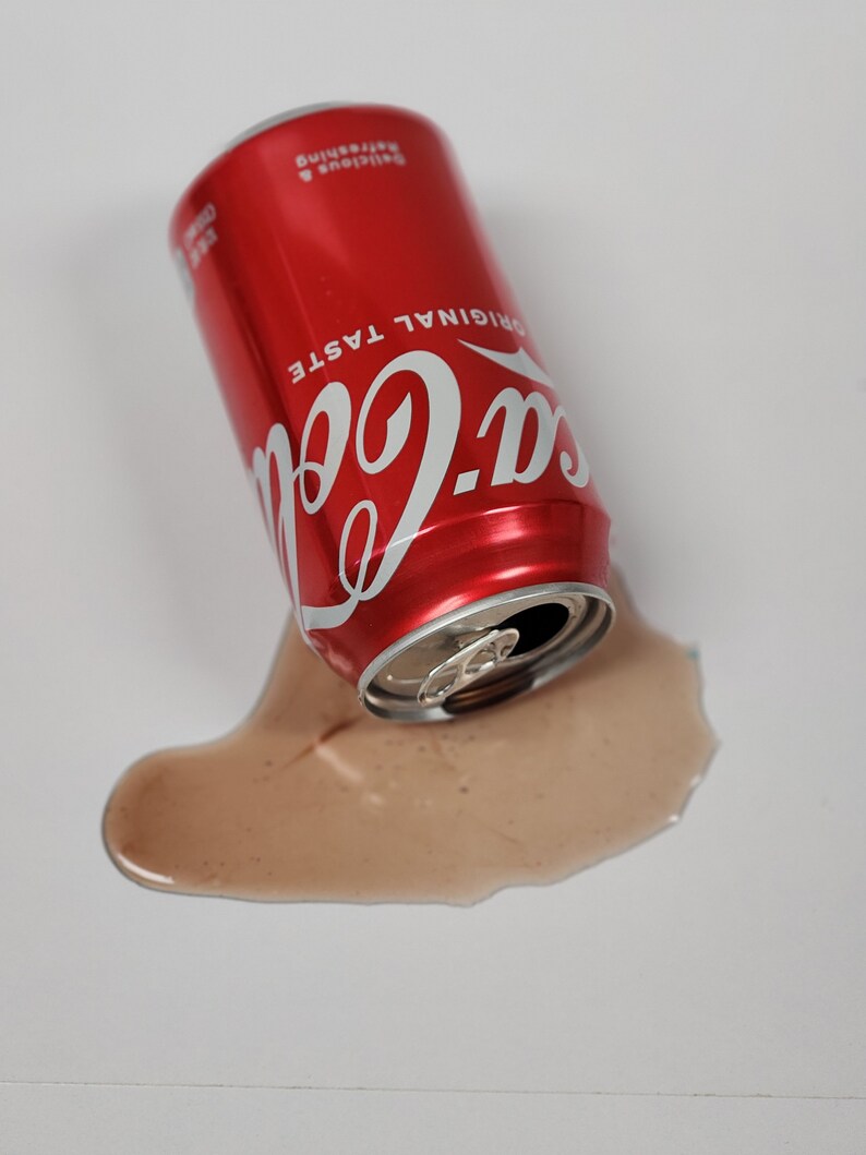 May include: A red can of Coca-Cola with the words "Original Taste" and "Delicious & Refreshing" printed on the side. The can is lying on its side and has spilled a brown liquid onto a white surface.