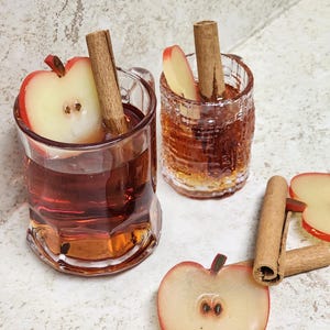 May include: Two clear glass mugs and a small glass, each filled with a reddish-brown liquid. One mug has a handle and a half apple and cinnamon stick. The other has a cinnamon stick and apple slice. A half apple and cinnamon sticks are on the surface.