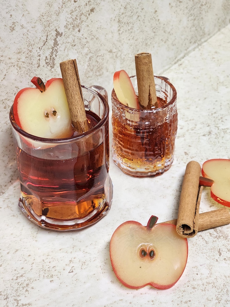 May include: Two clear glass mugs and a small glass, each filled with a reddish-brown liquid. One mug has a handle and a half apple and cinnamon stick. The other has a cinnamon stick and apple slice. A half apple and cinnamon sticks are on the surface.