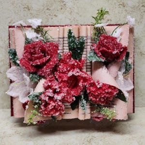 May include: A vintage book with red roses and white flowers emerging from the pages. The book is covered in faux snow.