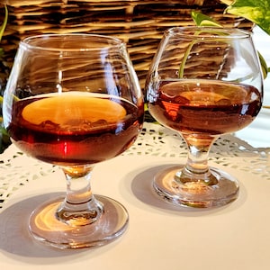May include: Two clear glass stemmed glasses filled with a dark amber liquid, sitting on a white lace doily.
