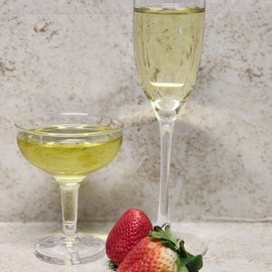Può includere: Due flûte da champagne e un bicchiere coupé riempiti con un liquido dorato, probabilmente champagne. Due fragole rosse mature con foglie verdi sono davanti ai bicchieri. I bicchieri sono su una superficie chiara.